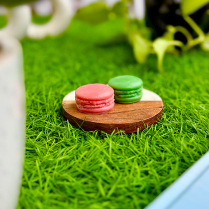 Two macarons, one pink and one green, on a wooden coaster with grass-like texture in the background.