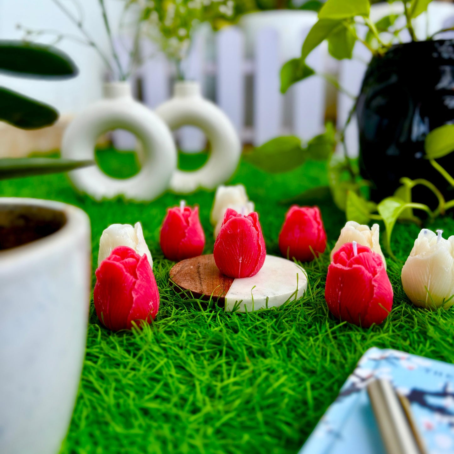 Decorative red and white flower-shaped candles on a grass-like surface with plants and a white fence in the background.